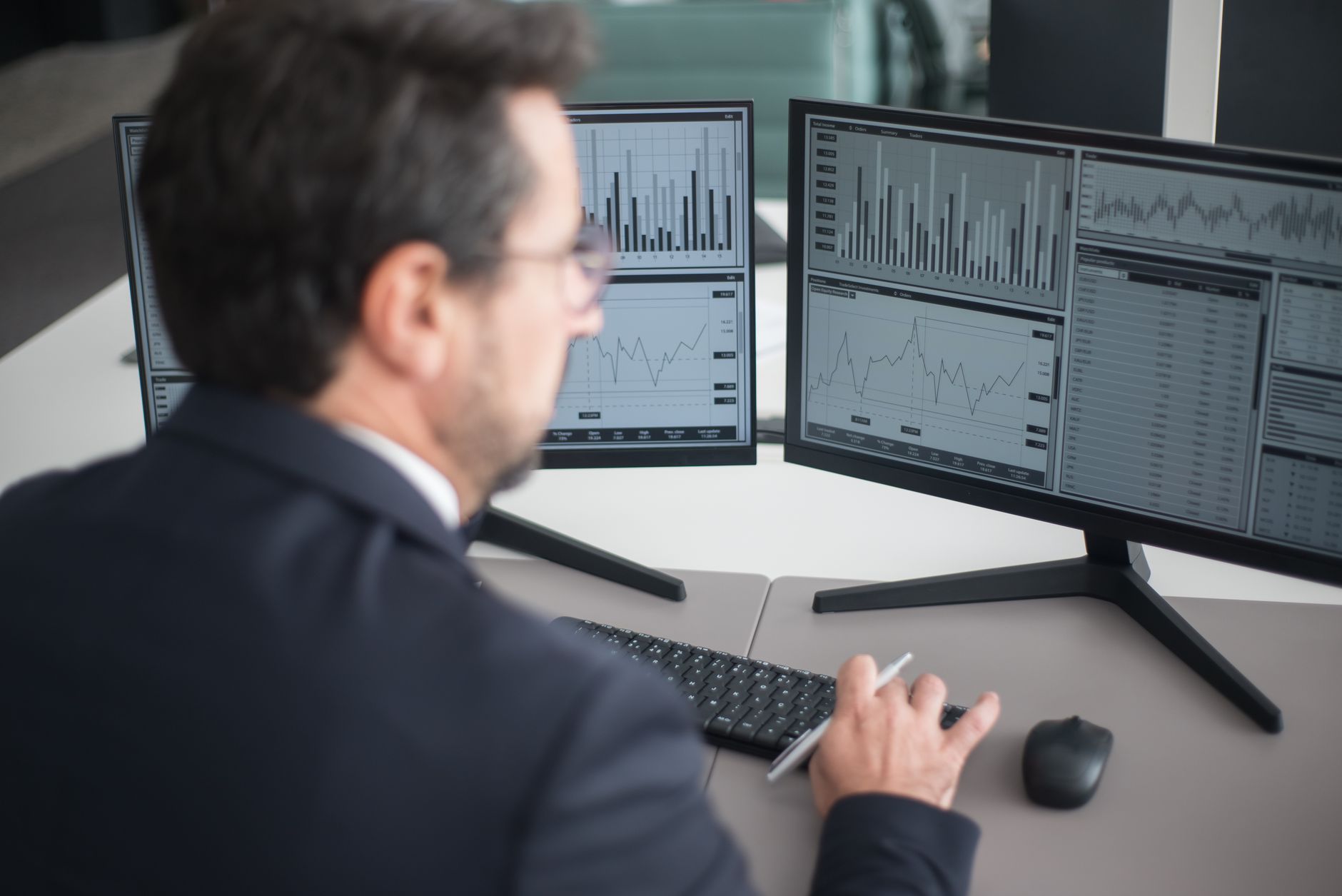 A focused South Korean export sales manager sitting at a desk with two monitors, comparing a printout datasheet with on-screen CRM data under warm office lighting.