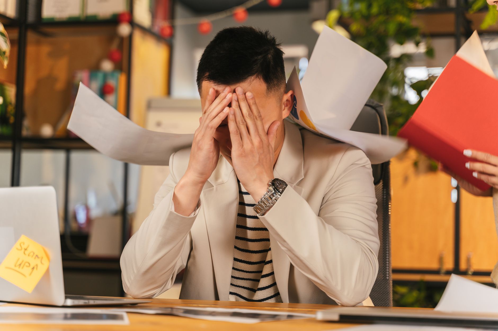 Image Generation Prompt: The back view of an office worker in their 30s clutching their head while looking at a pile of crumpled foreign business cards stacked high on an office desk.